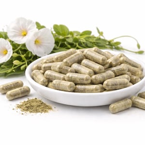 May include: A white bowl filled with numerous green capsules, alongside a small pile of green powder. White flowers with yellow centers and green leaves are arranged in the background, all set against a white backdrop.