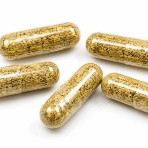May include: Five clear gelatin capsules filled with a coarse, granular, light brown herbal powder. The capsules are arranged in a scattered formation, showcasing the textured, earthy-toned contents visible through the transparent outer shell.
