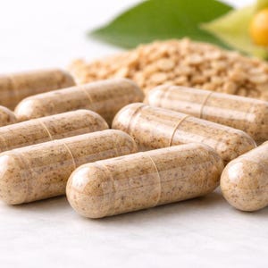 May include: Close-up of several light brown capsules, possibly supplements, arranged on a white surface. The capsules are cylindrical with a slightly textured appearance. In the background, there are small, tan granules and a green leaf.