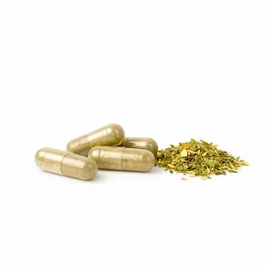 May include: Four light brown capsules are positioned near a pile of dried green herbs on a white background. The capsules are translucent, showing the herbal contents. The herbs are a mix of green and yellow, with a textured appearance.