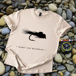 May include: A beige t-shirt featuring a black silhouette of a Hare's Ear Beadhead fly fishing lure. The text "- HARES EAR BEADHEAD -" is printed below the graphic. A fishing reel is visible in the background.