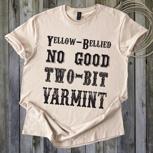 Puede incluir: Camiseta beige con la frase "YELLOW-BELLIED NO GOOD TWO-BIT VARMINT" en negro y fuente estilizada. La camiseta está doblada sobre una superficie de madera, con una cuerda enrollada en el fondo.