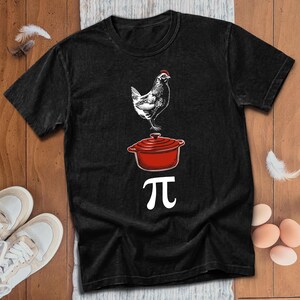 May include: A black short-sleeve t-shirt featuring a graphic print of a white chicken, a red lidded pot, and the white mathematical symbol for pi arranged vertically. The design creates a visual pun for chicken pot pie on a dark fabric.