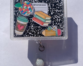 May include: A clear, glitter-edged badge reel featuring a composition book design with colorful school-themed charms. Charms include an apple, books, coffee cup, and pencil. The reel has a clear plastic strap and a silver-colored clip.