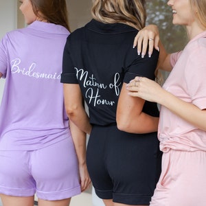 May include: Three women wearing matching pajama sets with the words "Bridesmaid", "Maid of Honor", and "Bride" on the back. The pajamas are in shades of purple, black, and pink.
