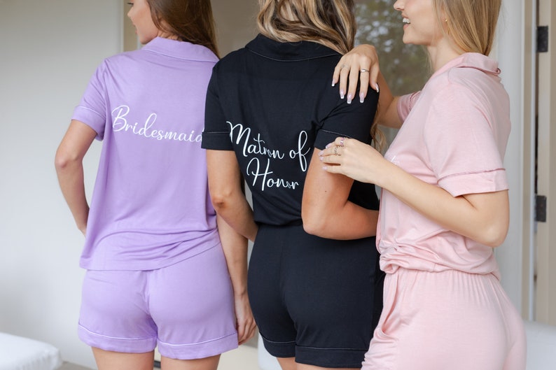 May include: Three women wearing matching pajama sets with the words "Bridesmaid", "Maid of Honor", and "Bride" on the back. The pajamas are in shades of purple, black, and pink.