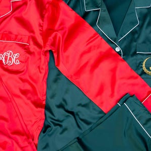 May include: Red and teal satin pajama sets. The red set has white piping and an embroidered monogram. The teal set has white piping and an embroidered initial within a gold wreath. Both sets have a button-down top and pants.