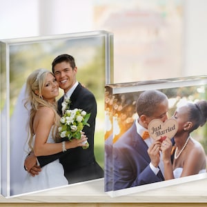 May include: Two clear acrylic photo blocks displaying wedding photos. One shows a smiling couple, the other a couple kissing behind a heart-shaped sign that reads "Just Married". The blocks are on a light-colored surface.