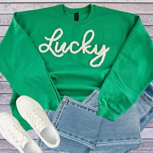 May include: A green sweatshirt with the word "Lucky" in white, paired with blue jeans and white sneakers. The sweatshirt is a vibrant green color, and the word "Lucky" is in a cursive font.