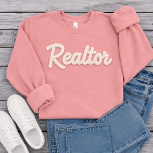 May include: A dusty pink crewneck sweatshirt featuring the word Realtor in white, textured, fuzzy chenille-style lettering across the chest. The garment is paired with blue denim jeans and a pair of white low-top sneakers with laces.