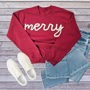 May include: A burgundy sweatshirt with the word "merry" in white script, paired with light blue jeans and white sneakers. The casual outfit is arranged on a wooden surface, suggesting a lifestyle or fashion theme.
