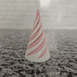 May include: A white and pink striped cone-shaped object. The object has a spiral design with alternating white and pink sections. The cone is standing upright on a speckled surface.