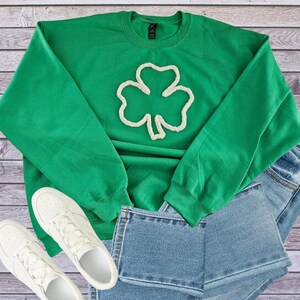 May include: A green sweatshirt with a white shamrock design, paired with denim shorts and white sneakers. The sweatshirt is a vibrant green color, and the shamrock is a prominent feature. The casual outfit is laid out on a wooden surface.