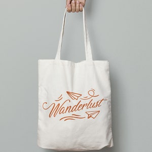May include: Cream-colored tote bag with the word "Wanderlust" in a stylized orange script, accompanied by paper airplane illustrations. The bag is held by a hand against a gray background. The tote bag has long straps.