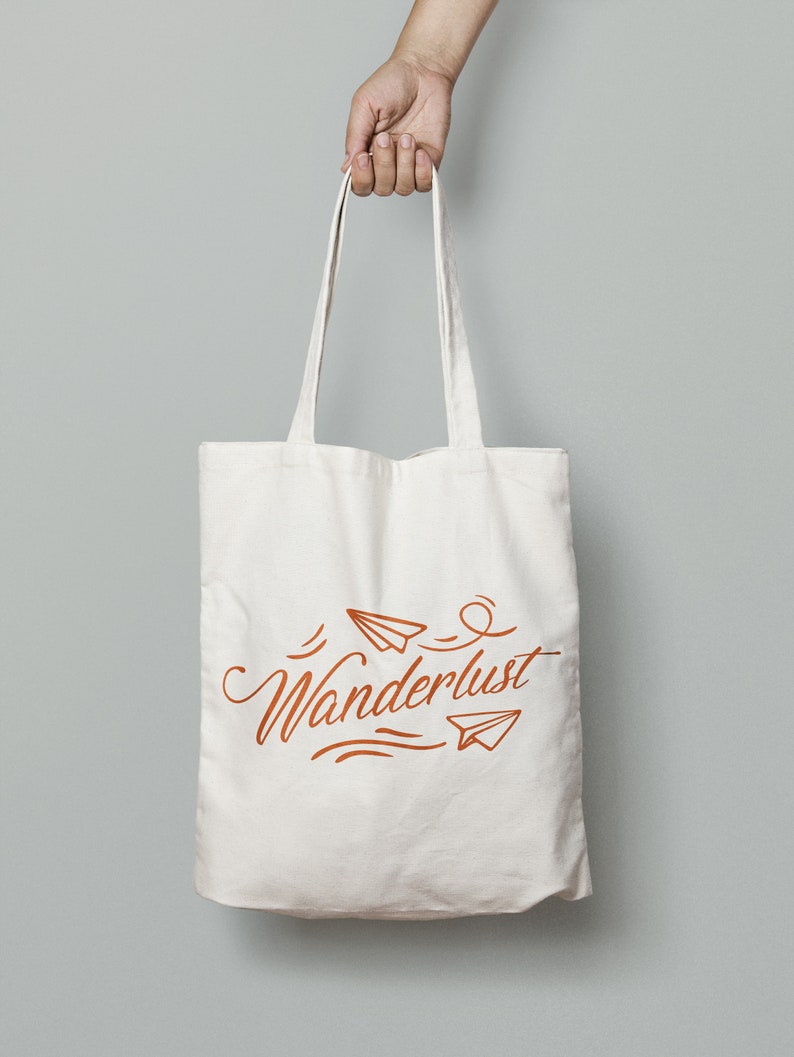 May include: Cream-colored tote bag with the word "Wanderlust" in a stylized orange script, accompanied by paper airplane illustrations. The bag is held by a hand against a gray background. The tote bag has long straps.