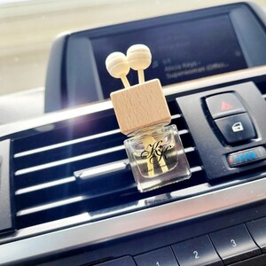 May include: A wooden car air freshener with a clear glass bottle filled with scented oil. The bottle has a wooden cap with two wooden balls on top. The air freshener is placed in the air vent of a car.