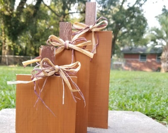 May include: Three wooden pumpkins of varying sizes, painted in a warm brown color. Each pumpkin is decorated with a brown and purple ribbon bow.