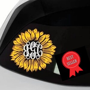 May include: A white car with a black rear window. A yellow sunflower with a white monogram in the center is on the window. A red and white best seller badge is on the window.