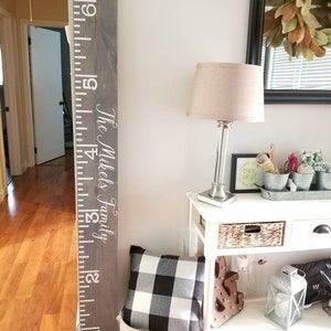 May include: A gray wooden growth chart with white numbers and the text "The Mikels Family". The chart is mounted on a white wall next to a white table with a lamp, a pillow, and other decor.