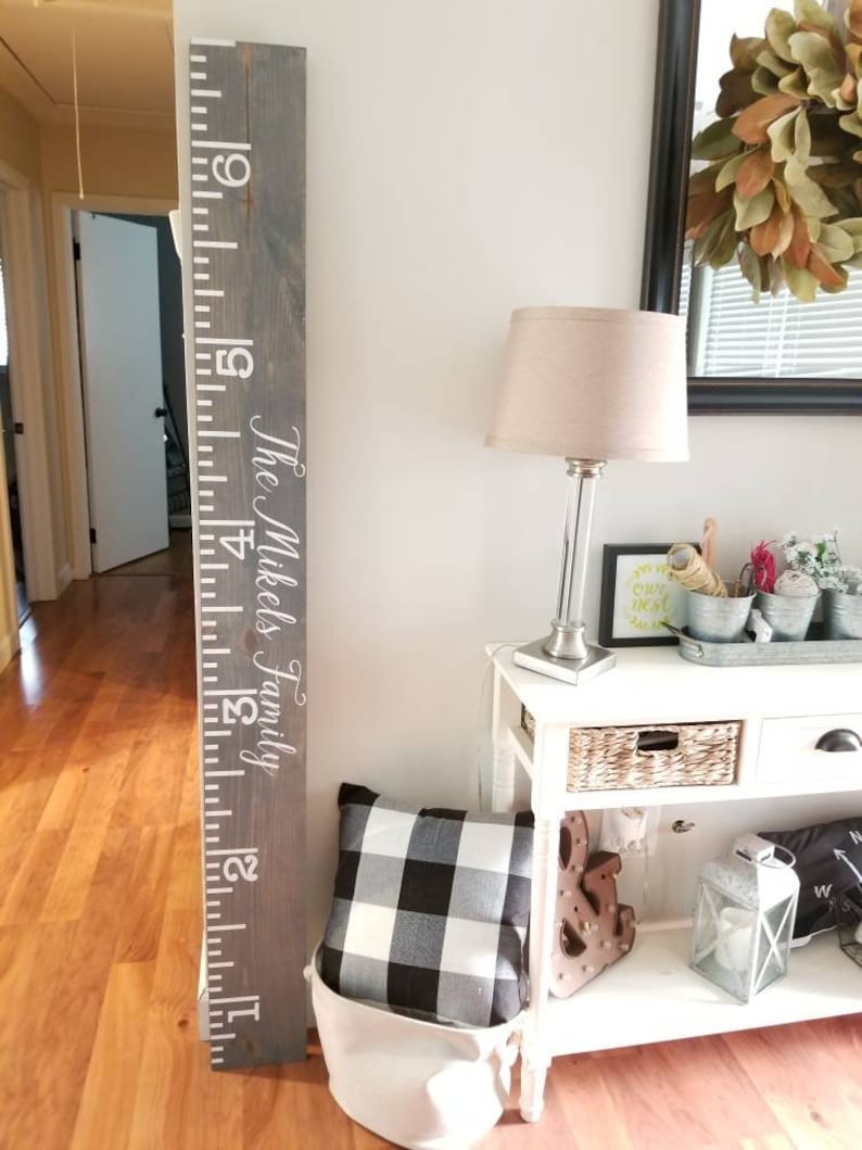May include: A gray wooden growth chart with white numbers and the text "The Mikels Family". The chart is mounted on a white wall next to a white table with a lamp, a pillow, and other decor.