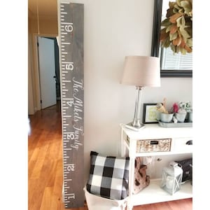 May include: A gray wooden growth chart with white numbers and the text "The Mikels Family" in white. The chart is mounted on a wall next to a white table with a lamp and decorative items.