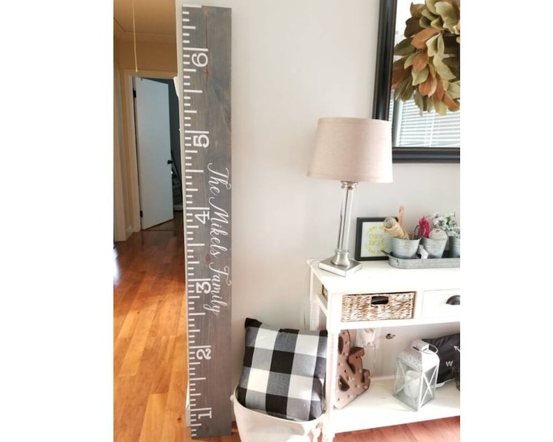 May include: A gray wooden growth chart with white numbers and the text "The Mikels Family" in white. The chart is mounted on a wall next to a white table with a lamp and decorative items.
