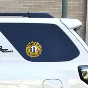 K&ouml;nnte beinhalten: Ein wei&szlig;er SUV mit einem gelben Sonnenblumen-Aufkleber auf der Heckscheibe. Die Sonnenblume hat einen wei&szlig;en Kreis in der Mitte mit den Buchstaben "AFA" in Blau.