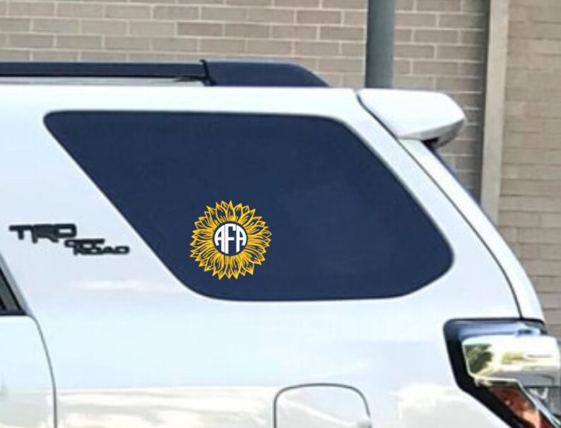 K&ouml;nnte beinhalten: Ein wei&szlig;er SUV mit einem gelben Sonnenblumen-Aufkleber auf der Heckscheibe. Die Sonnenblume hat einen wei&szlig;en Kreis in der Mitte mit den Buchstaben "AFA" in Blau.