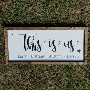 Könnte beinhalten: Ein Holzschild mit dem Text "This is us" in einer kursiven Schrift. Unter dem Text stehen die Namen Larry, Brittany, Autumn und Karsyn.