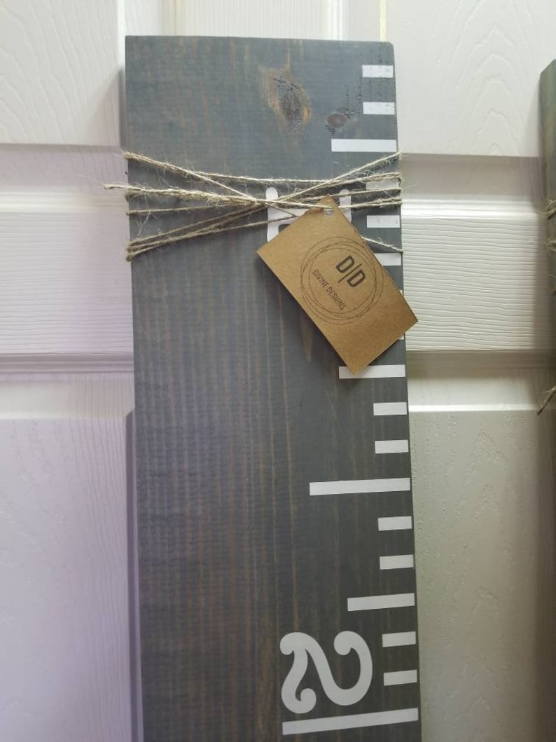 May include: A gray wooden growth chart with white markings and a white number "2" at the bottom. A brown tag with the text "D&D Divine Designs" is tied to the chart with twine.