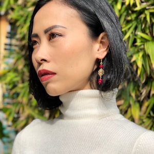 May include: A woman with short black hair wearing a white turtleneck sweater and gold earrings with red beads.