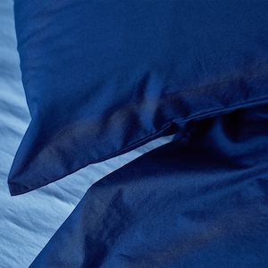 May include: A deep navy blue pillowcase resting on top of a matching navy blue duvet cover. The fabric has a smooth, matte texture with visible seams along the edges of the pillowcase, creating a clean and uniform appearance in the bedding set.