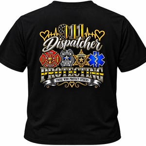 Pode incluir: Uma t-shirt preta de manga curta com um gráfico nas costas contendo o texto 911 Dispatcher, Protecting e Those Who Protect Others. O design inclui ícones de bombeiros, polícia, xerife e serviços médicos com detalhes em dourado.