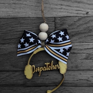 May include: Gold-colored wooden ornament shaped like a headset with the word "Dispatcher" written on it. The ornament is decorated with a black and white bow with gold stripes and white wooden beads.