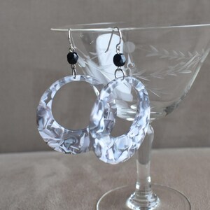 May include: A pair of dangle earrings featuring large, circular, white and grey marbled resin hoops. Each earring is suspended from a silver-toned metal hook and accented with a single, small, round, black faceted bead positioned above the hoop.