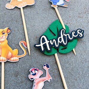 May include: A set of five cake toppers featuring characters from the Lion King movie. The toppers include Simba, Timon, Pumbaa, Zazu, and a green leaf with the name "Andres" written on it.