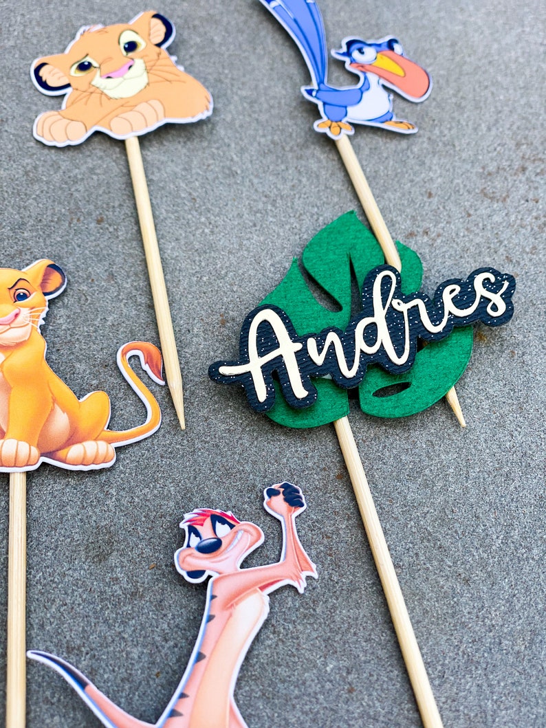 May include: A set of five cake toppers featuring characters from the Lion King movie. The toppers include Simba, Timon, Pumbaa, Zazu, and a green leaf with the name "Andres" written on it.