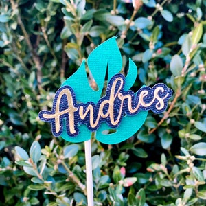 May include: A green leaf-shaped cake topper with the name "Andres" written in gold glitter letters on a black background.