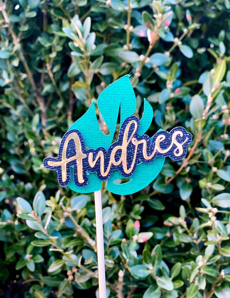 May include: A green leaf-shaped cake topper with the name "Andres" written in gold glitter letters on a black background.