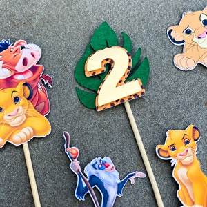 May include: A set of six cake toppers featuring characters from the Lion King movie. The toppers include Simba, Nala, Pumbaa, Timon, Rafiki, and a green leaf with the number 2 on it. The toppers are perfect for a Lion King themed birthday party.