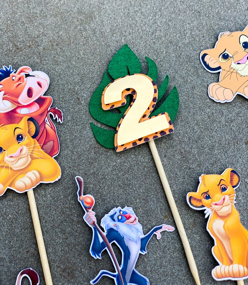 May include: A set of six cake toppers featuring characters from the Lion King movie. The toppers include Simba, Nala, Pumbaa, Timon, Rafiki, and a green leaf with the number 2 on it. The toppers are perfect for a Lion King themed birthday party.