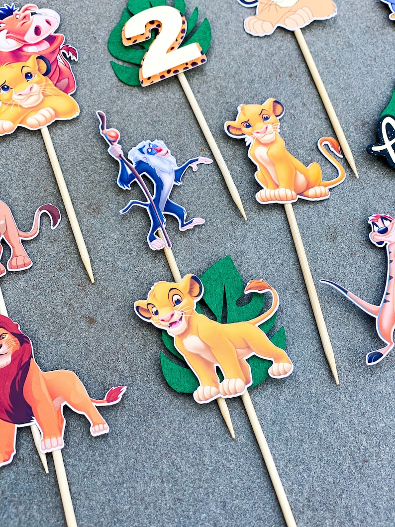 May include: A set of 10 Lion King themed cupcake toppers. The toppers feature Simba, Mufasa, Pumbaa, Timon, and Rafiki. The toppers are printed on cardstock and attached to wooden skewers.
