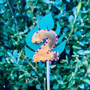 May include: A gold number two cake topper with a purple and orange leopard print design. The number two is on a green leaf with a white stick.