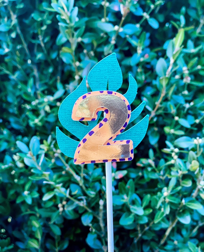 May include: A gold number two cake topper with a purple and orange leopard print design. The number two is on a green leaf with a white stick.