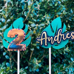 May include: Two cake toppers featuring a green monstera leaf design. One topper has a gold number 2 on the leaf and the other has the name "Andres" in black with gold glitter on the leaf.
