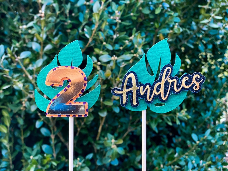 May include: Two cake toppers featuring a green monstera leaf design. One topper has a gold number 2 on the leaf and the other has the name "Andres" in black with gold glitter on the leaf.