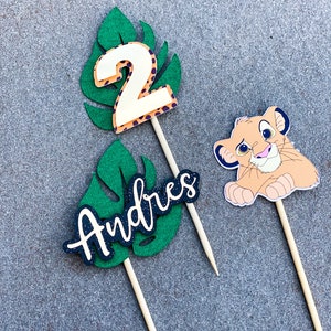 May include: Three cake toppers featuring a green leaf design with a white number 2, the name "Andres" in black lettering, and a cartoon lion cub.