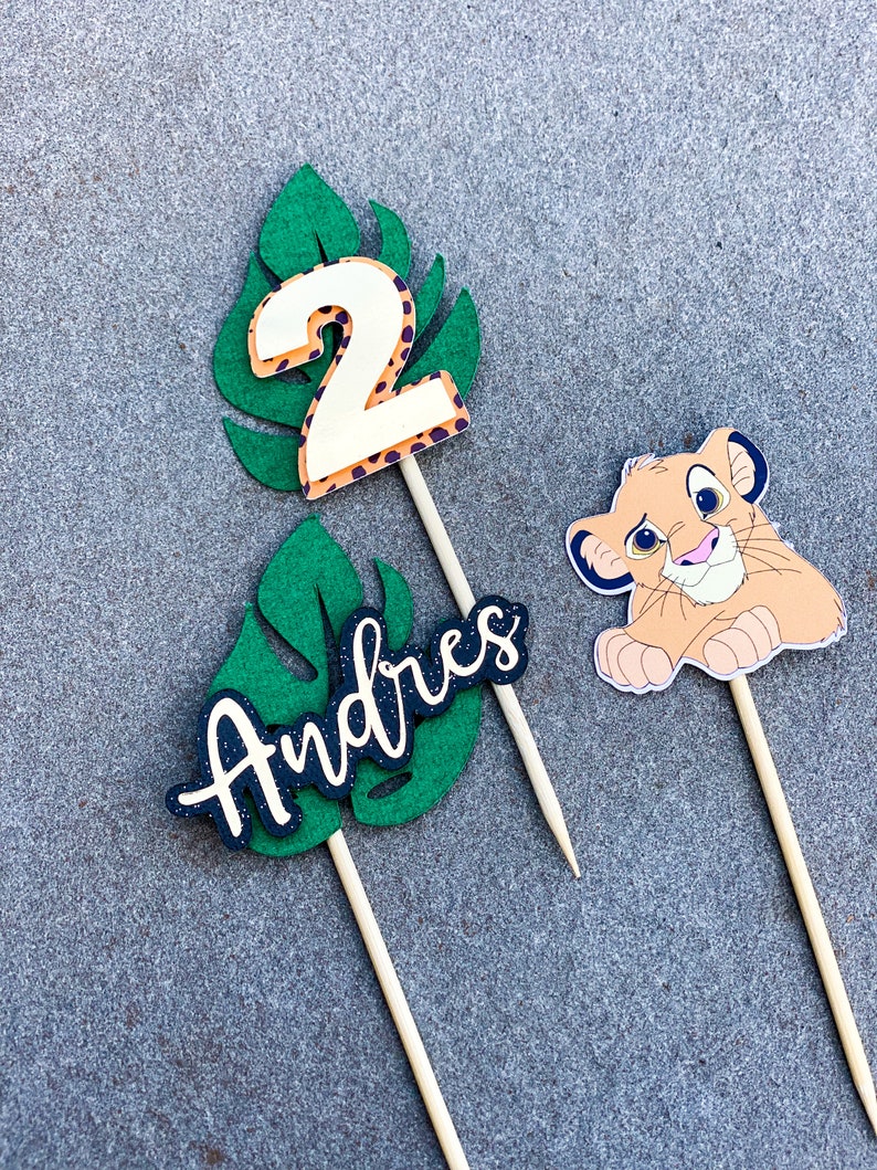 May include: Three cake toppers featuring a green leaf design with a white number 2, the name "Andres" in black lettering, and a cartoon lion cub.