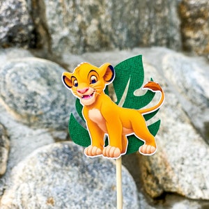 May include: A yellow lion cub cartoon character with a green leaf behind it. The character is on a wooden stick.