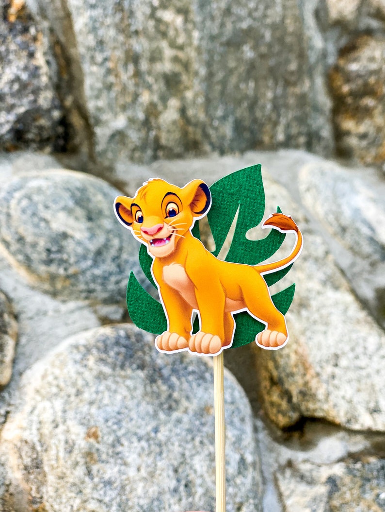 May include: A yellow lion cub cartoon character with a green leaf behind it. The character is on a wooden stick.